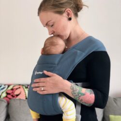 Kvinna bär spädbarn i en blå sele av märke Ergobaby